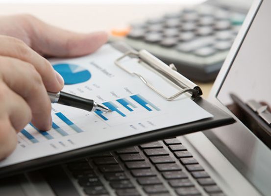 Profissional analisando gráficos financeiros em relatório impresso sobre notebook, representando os impactos da moeda funcional no SPED Contábil e na gestão tributária empresarial.
